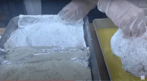Person breading food with flour and breadcrumbs. Gloved hands coating in flour before dipping in egg wash on baking tray.